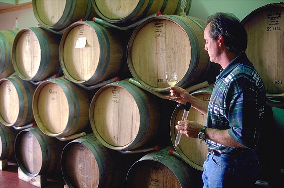 A man holds a wine glass in front of storage barrels