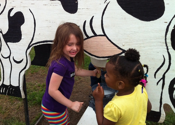 Two children participating in cow milking simulation with wooden cutout of a dairy cow