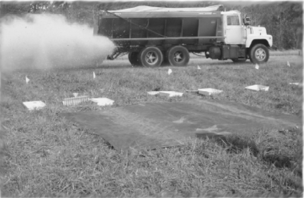 Black and white photo of truck with spreader in use near callibration setup with tarp, flags, collection containers etc.