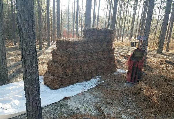 Empty baler next to several neatly stacked pine straw bales on a piece of plastic sheeting among a stand of trees.