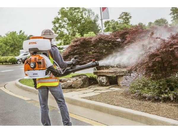 Worker stands near curb spraying small trees with backpack mistblower