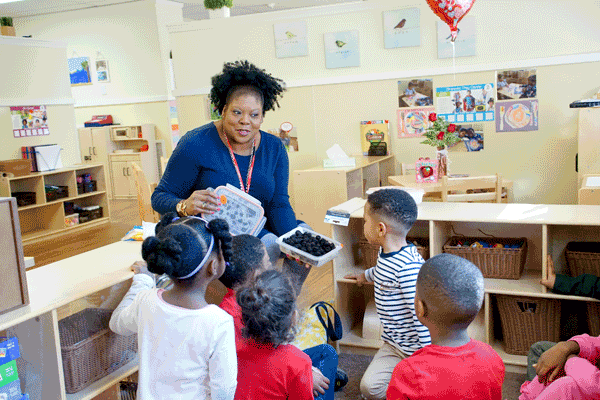 An adult offers a group of young children a container of fruit in a classroom.