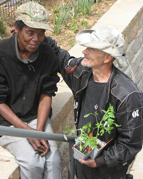 One garner has arm around other gardener's shoulder and holds a plant