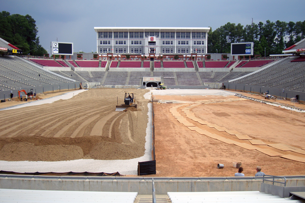 Laser-guided bulldozer spreads soil mix in stadium as fence protects sub-surface layers.