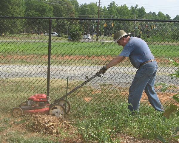 A gardener pushes a lawnmower near a fence