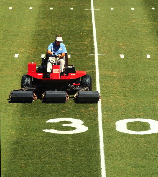 A person drives a reel mower along the 30-yard line of a football field.