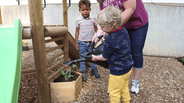 An adult helps young children use a watering can to water plants growing in a garden box next to a play set.