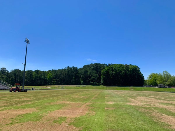 An athletic field that appears patchy with a tractor pulling a fraise mower in the distance.