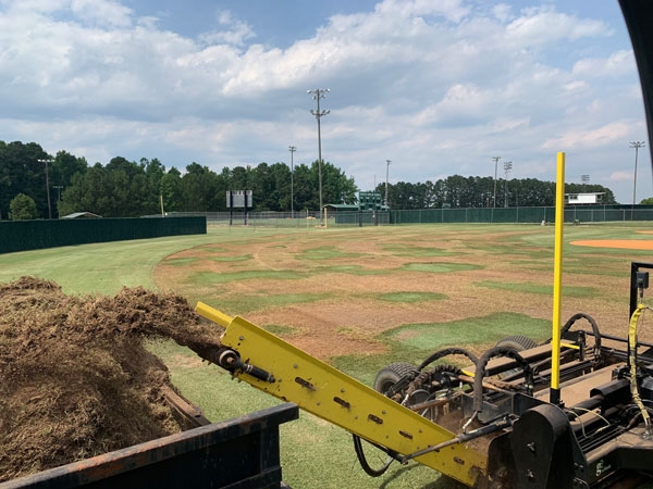 Debris being expelled to a dump trailer in the foreground with a patchy athletic field in the background.