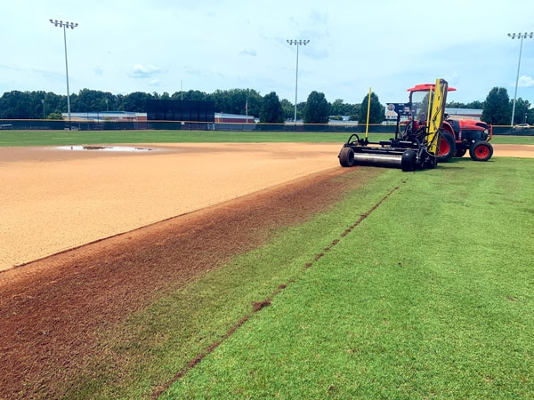 A tractor pulls a fraise mower along an athletic field, leaving a lip.