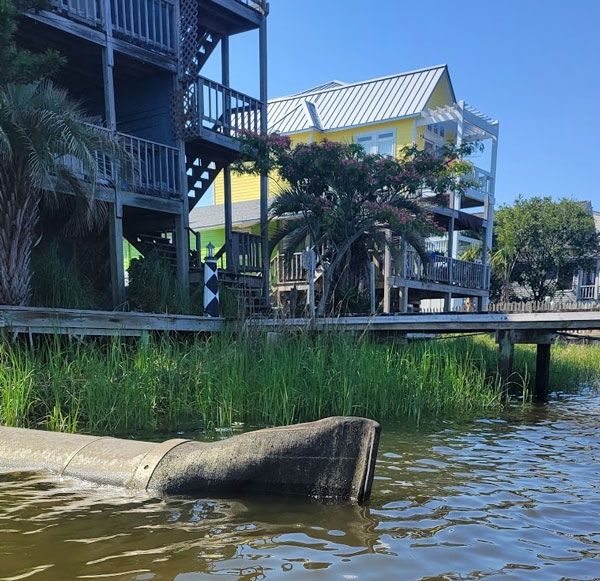 Photo: A black rubber duckbill check valve attached to a pipe coming out of the shore. The pipe is about half submerged. Tall grass and a dock behind pipe.