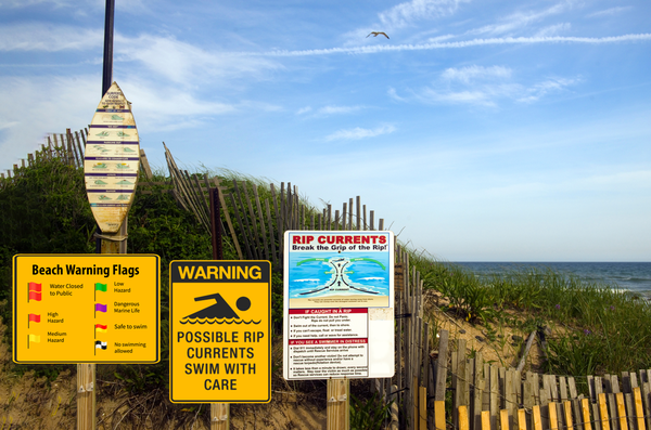 Signs on a sand dune showing beach warning flags and warning about rip currents.