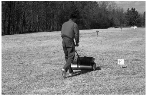 Black and white photo of someone pushing drop spreader with catch pan at starting line marked with small sign