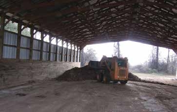 Interior of a large, open-sided barn with a metal roof and partial siding. A pile of dark brown poultry litter is visible on the ground and a small skid steer next to the pile