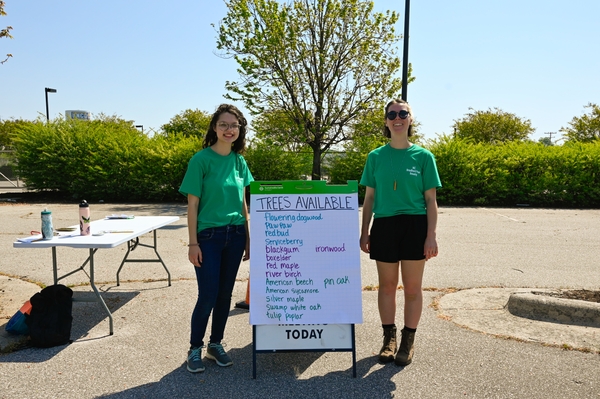 Two people stand next to a list of trees written on large sandwich board