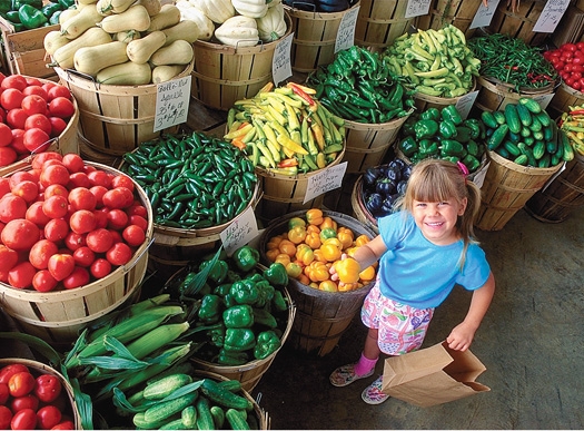 A child holds a bell pepper and smiles in front of baskets of vegetables at a produce stand