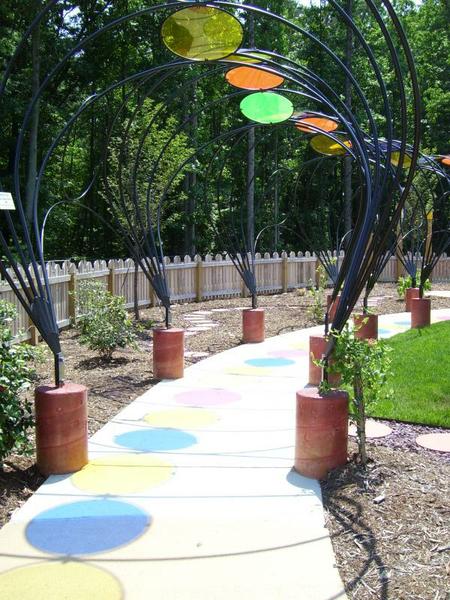 sculpture overhangs sidewalk and light passes through colored circles on sculpture to create shadow pattern on the path